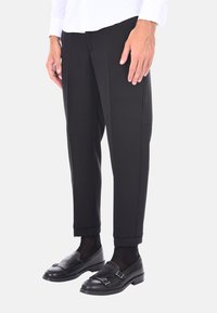 Pantaloni neri su misura con una texture liscia, caratterizzati da una forma affusolata e dettagli sulle cuciture laterali, abbinati a mocassini neri e calze scure.