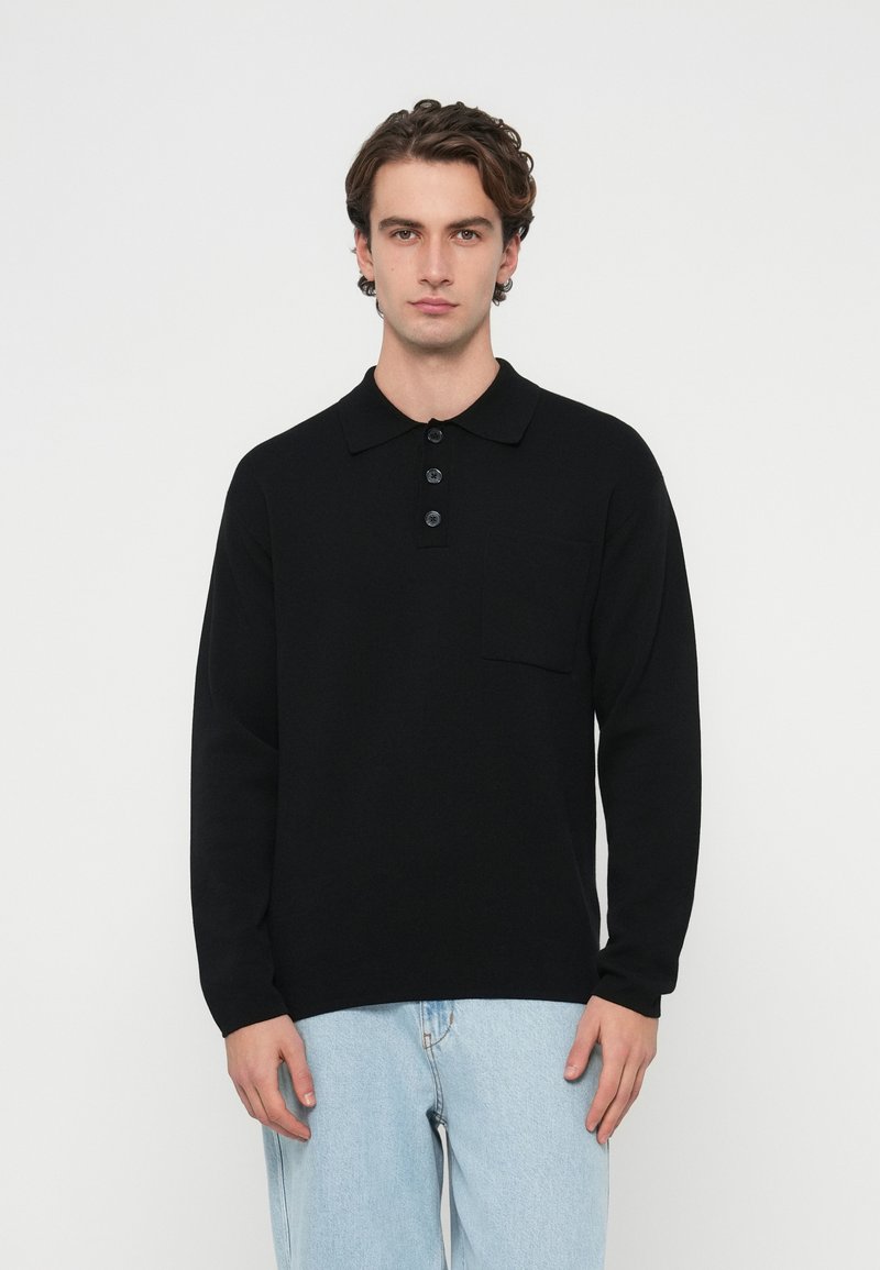 Czarna koszulka polo z długim rękawem, kołnierzykiem, listwą na pięć guzików oraz kieszenią z przodu; w zestawieniu z jasnoniebieskimi jeansami. Gładka tekstura.