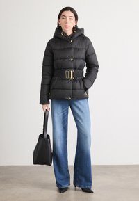 Zwarte pufferjack met een ceintuur in de taille, gouden hardware, capuchon en geborduurd ontwerp, gecombineerd met brede, blauwe jeans en een zwarte handtas.