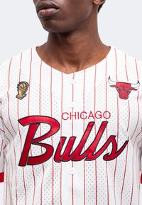 Νεαρός άνδρας φοράει μια λευκή φανέλα των Chicago Bulls με κόκκινες ρίγες, όνομα ομάδας, λογότυπο και ένα εμβληματικό έπαθλο πρωταθλήματος NBA.