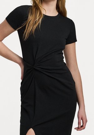 Femme portant une robe noire à manches courtes avec un détail nœud torsadé à la taille et une petite fente à l'avant.