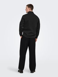 Homme debout, tourné vers l'arrière, portant un sweat-shirt noir, un pantalon noir ample et des chaussures noires, sur un fond clair uni.