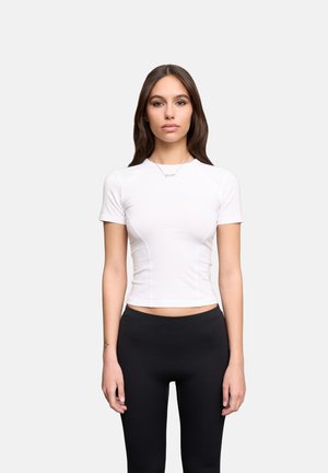 Jeune femme aux longs cheveux bruns, portant un haut blanc ajusté à manches courtes et des leggings noirs, debout devant un fond clair uni.