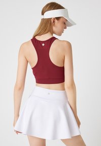 Burgundy racerback urheilullinen crop-top, yhdistettynä valkoiseen liehuvaan hameeseen. Topissa on istuva malli ja logotyyli takana.