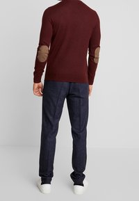 Pull bordeaux avec coudières marron, associé à un pantalon texturé foncé et des baskets blanches, vu de dos.
