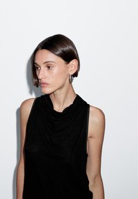 Top senza maniche nero con scollatura drappeggiata. Tessuto morbido e textured, design minimale. Modella che indossa grandi orecchini a cerchio d'argento e ha i capelli corti.