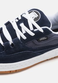 Tengerészkék velúr és hálós sportcipő fehér fűzőkkel, Vans logóval ellátott címkével és gumi talppal. Különböző textúrák és színek keveréke.