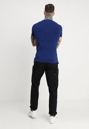 Homme tatoué portant un t-shirt bleu, un pantalon cargo noir et des baskets blanches, debout les bras croisés, de dos sur un fond uni.