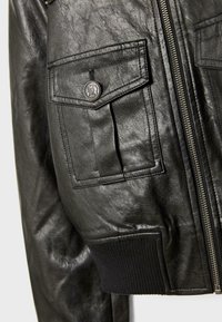 Crna kožna bomber jakna s patent zatvaračem, rebrastim manžetama i džepom na prsima s metalnim kopčanjem. Vidljiva teksturirana obrada.