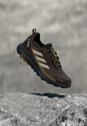 Brauner und schwarzer Adidas Terrex Trailrunning-Schuh mit weißen Streifen, über felsigem Gelände schwebend, der dynamische Nutzung im Freien zeigt.