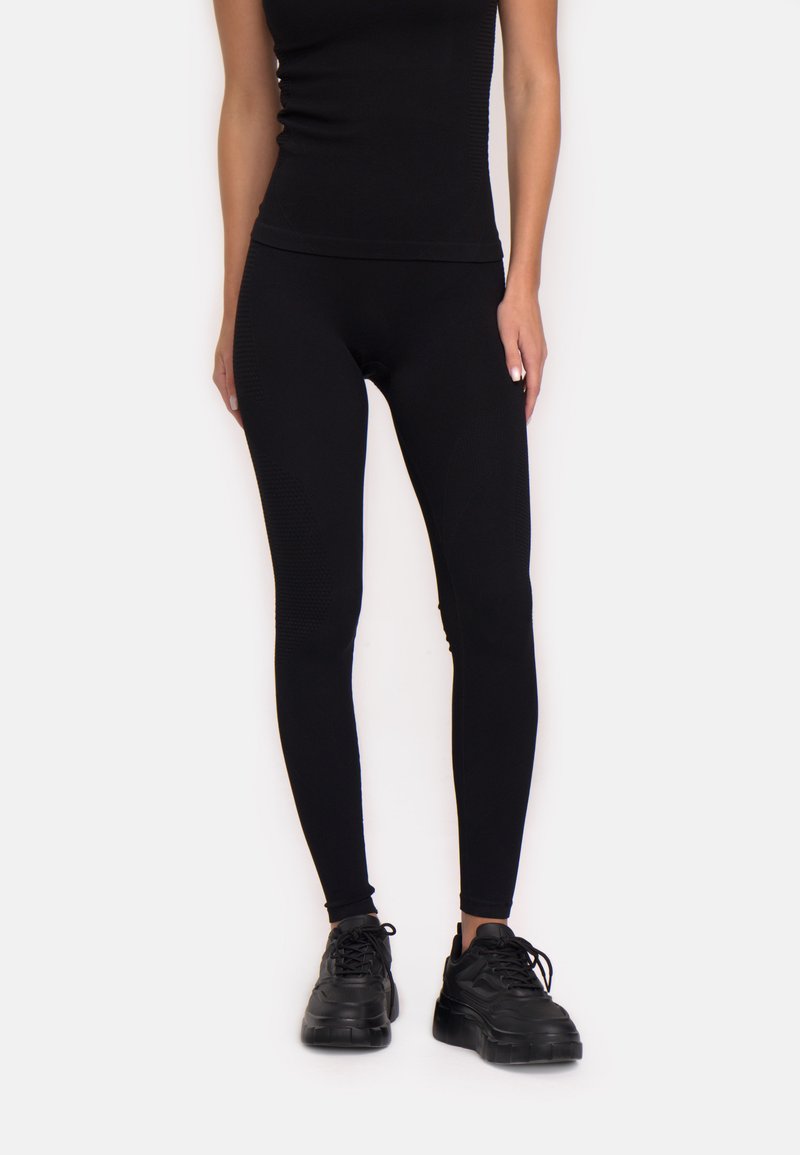 Kobieta ubrana w czarne dopasowane legginsy, czarny top na ramiączkach oraz masywne czarne sneakersy, stojąca na tle białej ściany.