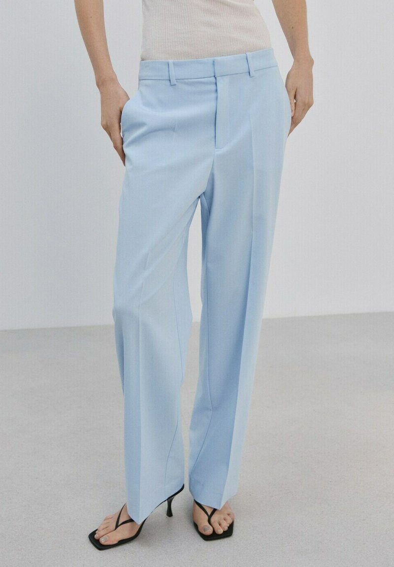 Frau trägt hellblaue, maßgeschneiderte, weit geschnittene Hosen, weißes, geripptes ärmelloses Oberteil und schwarze Sandalen mit dünnen Absätzen.