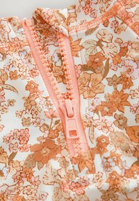 Tissu à motifs floraux dans des tons de pêche doux et de crème, avec une fermeture éclair rose centrale et un col froncé. Texture lisse et légère.