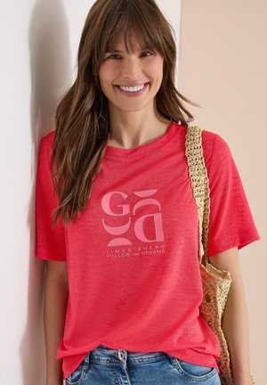 Smilende kvinde med langt brunt hår iført en rød grafisk T-shirt med teksten "Times Ahead Follow Your Dreams", blå jeans og bærer en beige flettet taske.