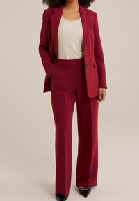 Bordowy garnitur z dopasowaną marynarką i szerokimi spodniami, w połączeniu z dopasowaną jasną bluzką. Gładki materiał i metalowe detale przy guzikach.