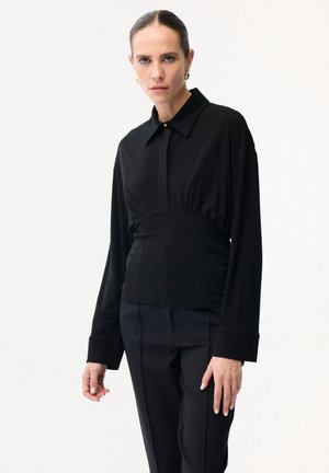 Femme aux cheveux foncés coiffés en chignon élégant, portant un chemisier noir ajusté à manches longues et un pantalon noir, posant devant un fond blanc.