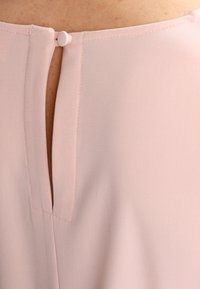 Blusa de color rosa claro con apertura en la espalda en forma de llave y cierre de botón, con una textura de tela suave y lisa. Diseño minimalista.
