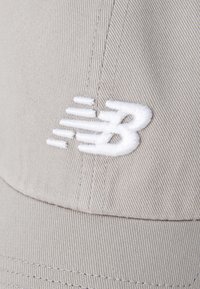 Helle graue Kappe mit strukturiertem Design, ausgestattet mit einem weißen gestickten Logo. Der Stoff hat eine glatte Textur und sichtbare Nähdetails.