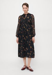 Svart blomstret kjole med lange transparente ermer, samlet midje og knappeåpning. Har flerfargede blomster mønstre og et knyting detalj ved halsen.