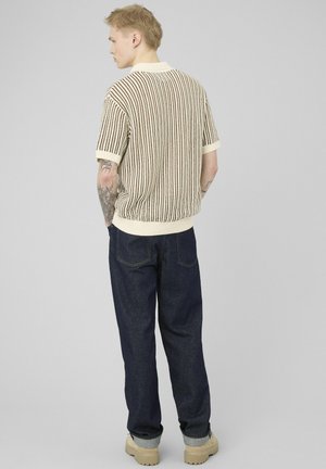 Homme tatoué portant un pull en maille beige et marron à rayures à manches courtes, un jean foncé retroussé et des chaussures beiges, vu de dos sur un fond gris.