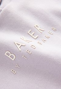 Lyse lilla stof med præget tekst, der lyder "BAKER by TED BAKER" i en metallisk finish, med en glat tekstur.