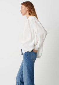 Blusa bianca a maniche lunghe con spalle arricciate, dal tessuto morbido e dal taglio rilassato, abbinata a jeans a gamba dritta blu chiaro.