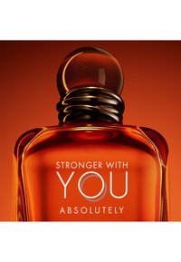 Amber glazen parfumfles met een ronde, zwarte dop. Witte tekst zegt "STRONGER WITH YOU ABSOLUTELY" op een levendige oranje achtergrond.