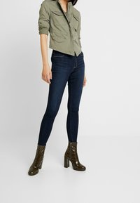 Olivgrön kort utility-jacka med flera fickor, parat med mörkblå skinny jeans och glansiga mörkbruna klackiga ankelstövlar.