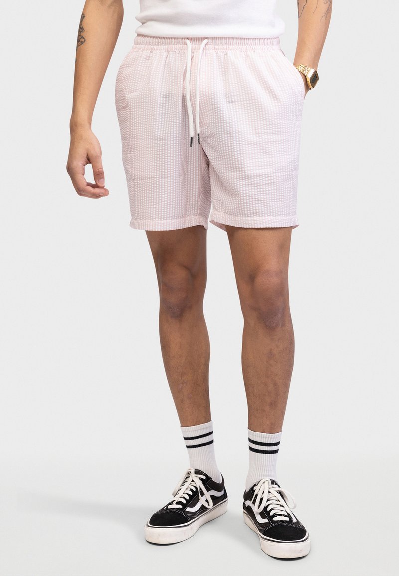 Άνδρας που φοράει ροζ ανοιχτό χρώμα υφασμένα σορτς, λευκές κάλτσες με μαύρες ρίγες, μαύρα και λευκά αθλητικά παπούτσια, λευκό πουκάμισο και χρυσό ρολόι χειρός.