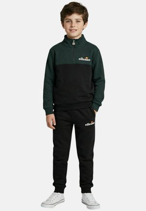 Garçon debout portant un survêtement ellesse noir et vert avec des baskets blanches, les mains dans les poches, souriant devant un fond uni.