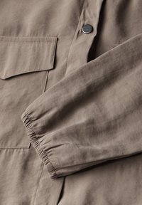 Veste marron clair présentant une texture douce, des poignets élastiques, une poche plate et un détail de bouton. Le matériau semble léger et lisse.