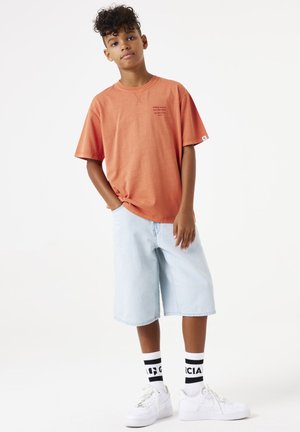 Oranje T-shirt met korte mouwen met een tekstontwerp, gecombineerd met lichtblauwe denimshorts en witte sneakers, met zwart-wit gestreepte sokken.