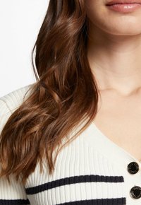 Krémový ribovaný cardigan s tmavě modrými pruhy, s výstřihem do V a třemi ozdobnými knoflíky na přední straně. Měkká textura, přiléhavý střih.