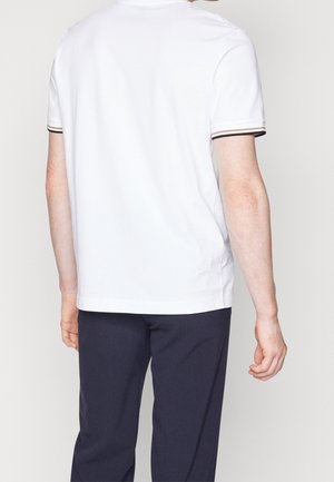 Man draagt een wit kortarmig hemd met gestreepte manchetten en donkere broek, gezien van achter tegen een effen achtergrond.