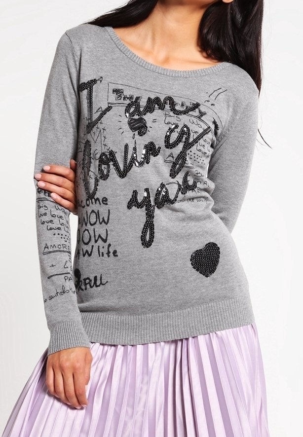 Femme portant un pull gris avec un texte noir « I am loving you » et des gribouillis, associé à une jupe plissée violet clair.