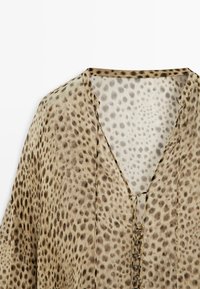 Beżowa bluzka w brązowe cętki leopardzie, z dekoltem w serek i małymi guzikami wzdłuż środka przodu.