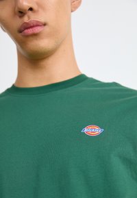 Zelené bavlnené tričko s okrúhlym, viacerými farbami logo Dickies na ľavej strane hrude. Dizajn s okrúhlym výstrihom a hladkou textúrou.