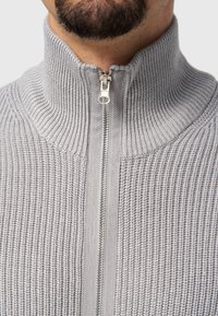 Suéter gris de cremallera con un cuello alto. Presenta una cremallera plateada y un tejido texturizado, mostrando un diseño casual y moderno.