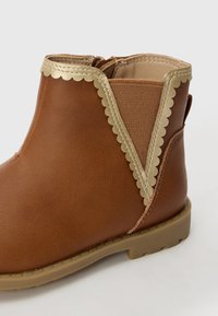 Botte cheville marron avec un bord doré créneau. Fabriquée en matériau synthétique lisse, elle présente un panneau élastique texturé et une semelle en caoutchouc.