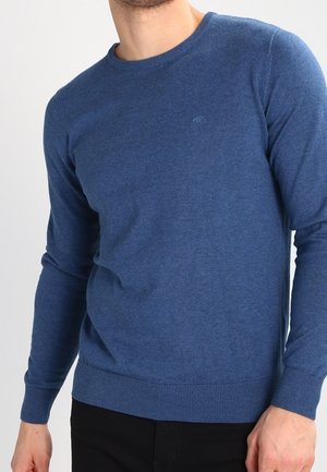 Mand iført en ensfarvet blå langærmet sweater med ribkanter ved ærmer og kant, stylet med sorte bukser, mod en hvid baggrund.