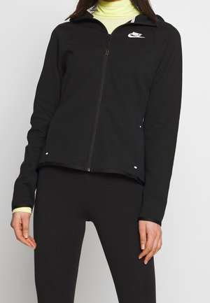 Femme portant une veste zippée noire Nike par-dessus un col roulé jaune, associée à un legging noir, debout devant un fond uni.