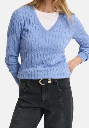 Femme portant un pull en maille torsadée bleu clair à col en V sur une chemise blanche, associé à un jean noir taille haute et une ceinture noire avec une boucle argentée.