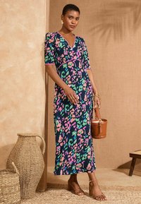 Maxi obleka v temno modri barvi z barvnim cvetličnim vzorcem, ki vključuje rožnate, zelene in modre odtenke. Kratki rokavi in V-izrez. Rjava torbica.