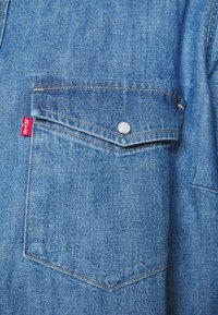 Giacca di denim di colore azzurro chiaro, dotata di una tasca sul petto con un bottone a scatto argentato e un'etichetta rossa sul lato.