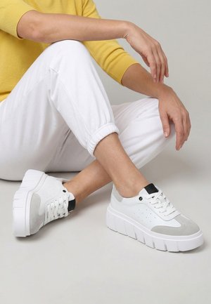 Person sitzt mit verschränkten Beinen, trägt weiße Plateausneaker, weiße Hose und ein gelbes Langarmoberteil vor einem einfarbigen grauen Hintergrund.