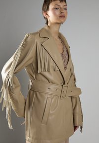 Beige Kunstledermantel mit Fransenverzierungen an den Schultern, breiten Revers, tailliertem Gürtel und seitlichen Taschen. Glatte Textur, moderner Schnitt.