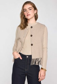 Gilet beige texturizzato con chiusura a bottone e orlo con frangia, abbinato a una maglietta beige chiaro e jeans in denim scuro. Accessori metallici semplici.