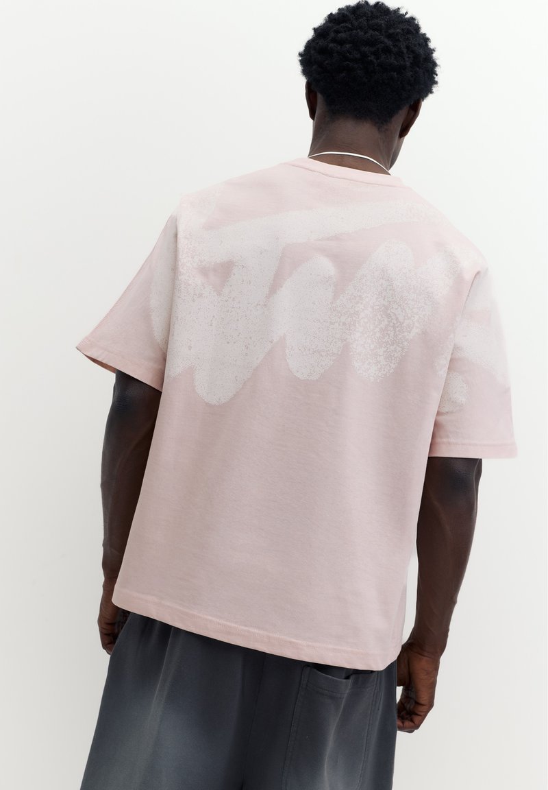 Homme aux cheveux courts et bouclés portant un t-shirt rose clair oversize avec un motif blanc délavé dans le dos et un pantalon sombre ample.