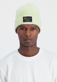 Világos zöld kötött sapka bordázott textúrával, fekete címkével, amelyen az Alpha Industries logója és szövege látható, egy modellen viselve.