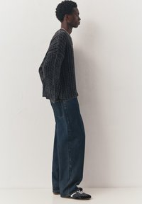 Junger Mann steht seitlich, trägt einen grauen Kabelstrickpullover, lockere dunkelblaue Jeans, schwarz-weiße Turnschuhe und eine silberne Kettenhalskette.